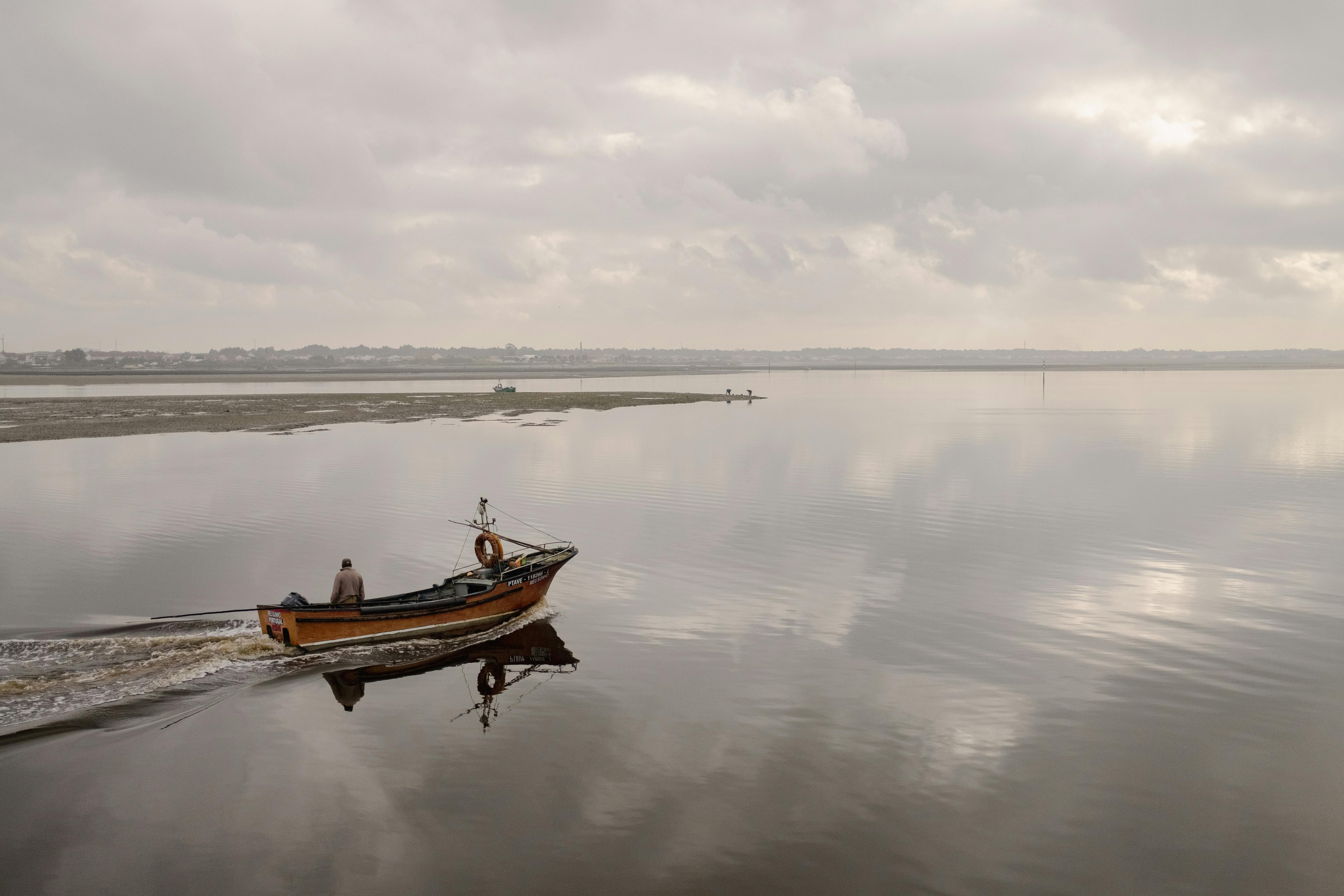 A lone boat sails quietly across the calm waters of Ria de Aveiro, Portugal under a cloudy sky.