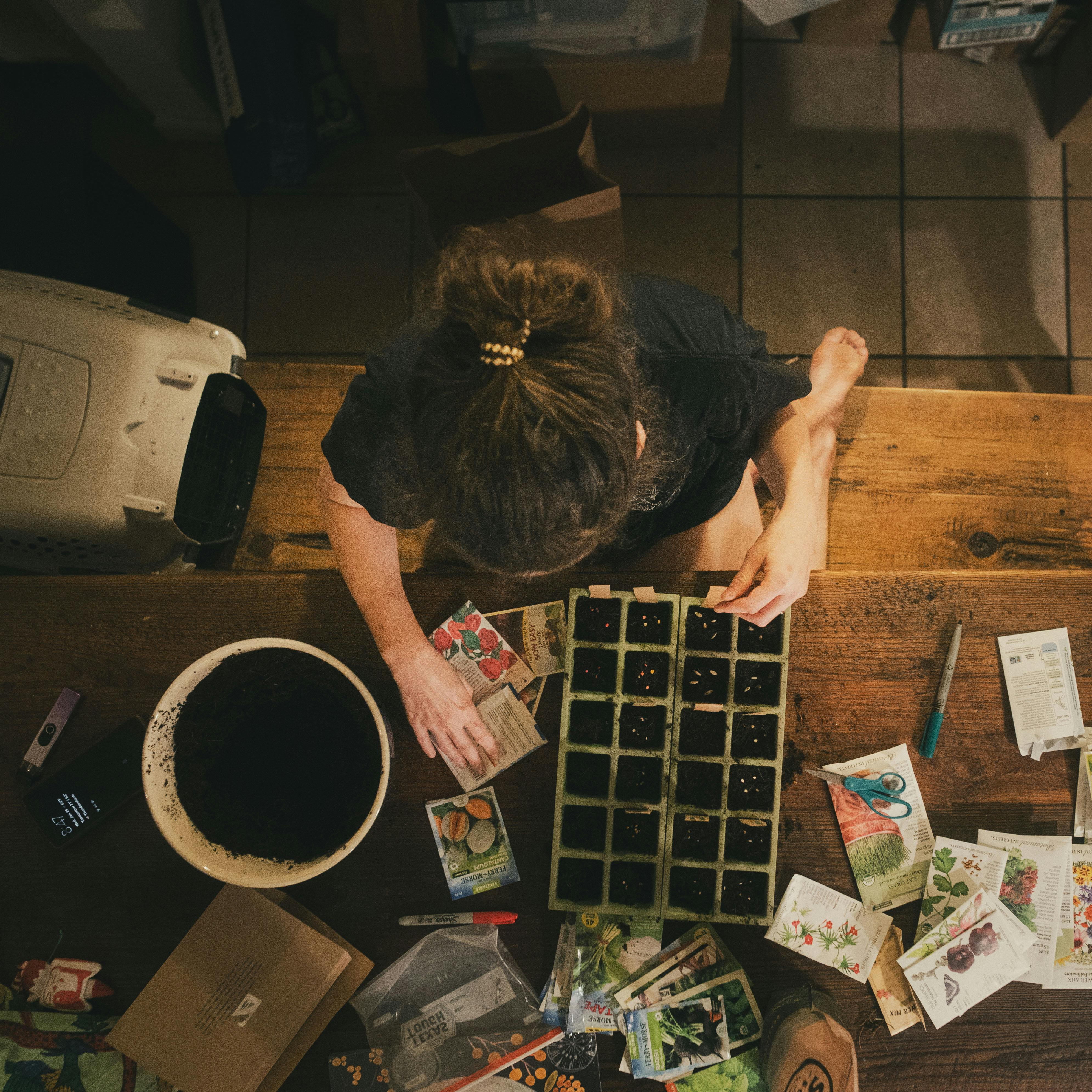 A person carefully plants seeds in containers on a wooden table indoors, focused on gardening.