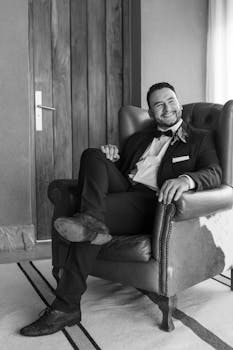 Black and white portrait of groom smiling while seated indoors in a formal attire.