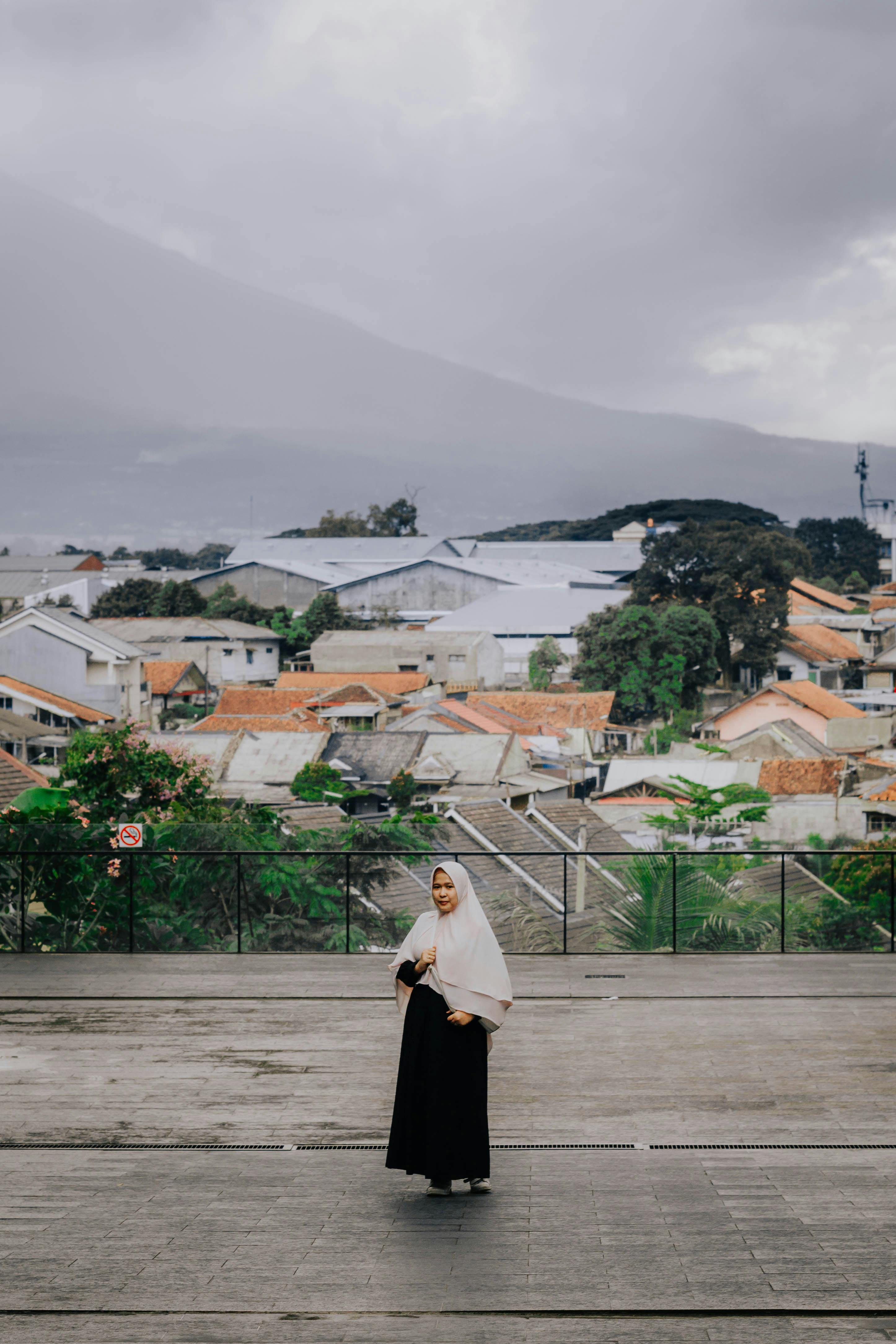 一位身着黑色连衣裙、头戴头巾的女子，正站在西爪哇的户外，身后是城市景观和连绵的山脉
