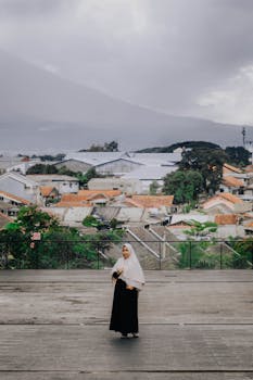 一位身着黑色连衣裙、头戴头巾的女子，正站在西爪哇的户外，身后是城市景观和连绵的山脉