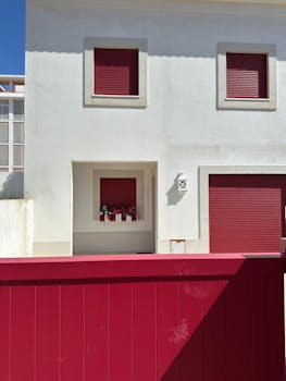 Minimalist white house exterior with striking red shutters and gate under bright sunlight.
