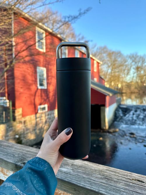 Free Person holds black water bottle in front of rustic red mill by the river. Stock Photo