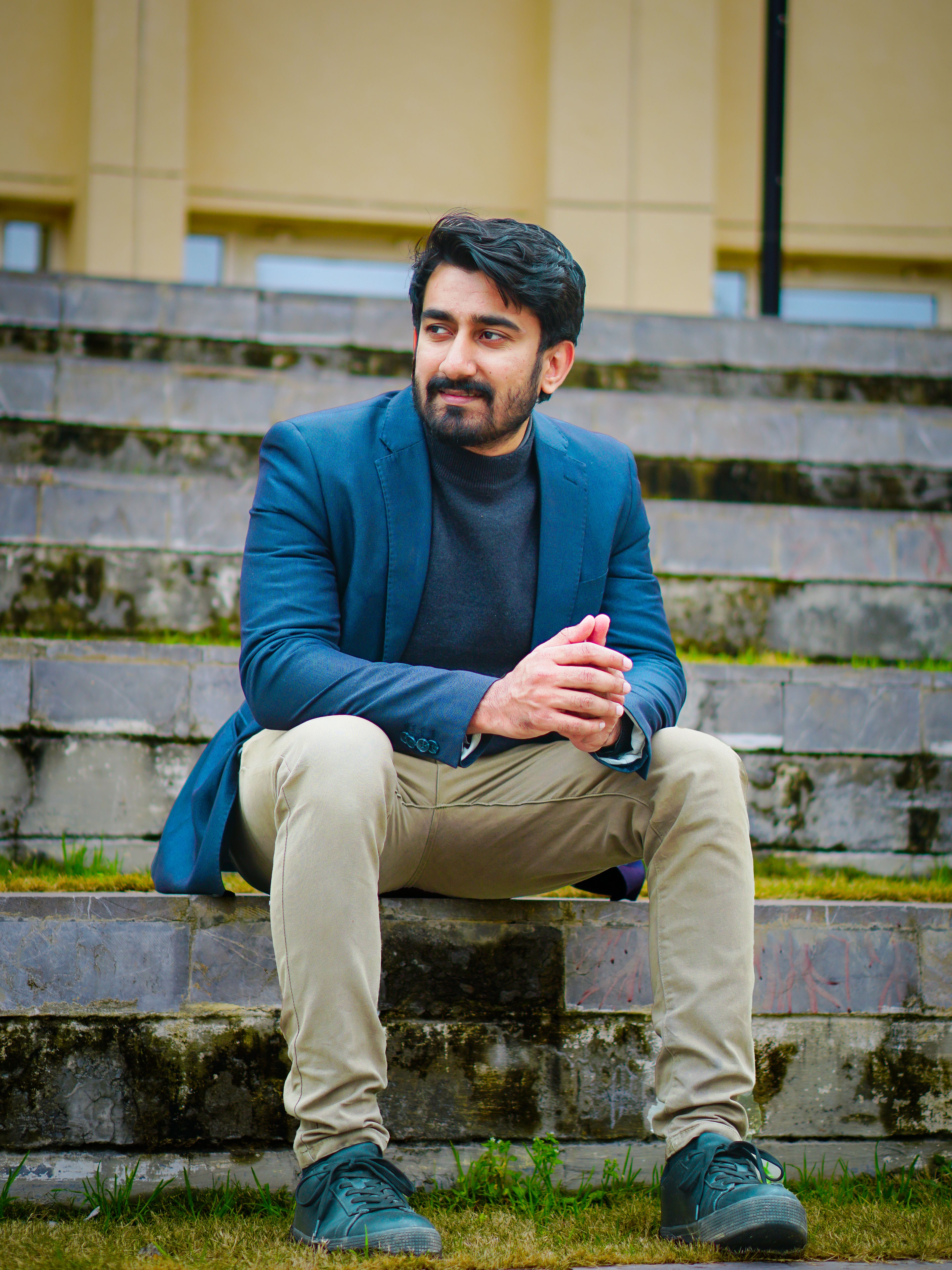 A man in a blue suit and beige pants sits on outdoor steps, exuding a relaxed style.