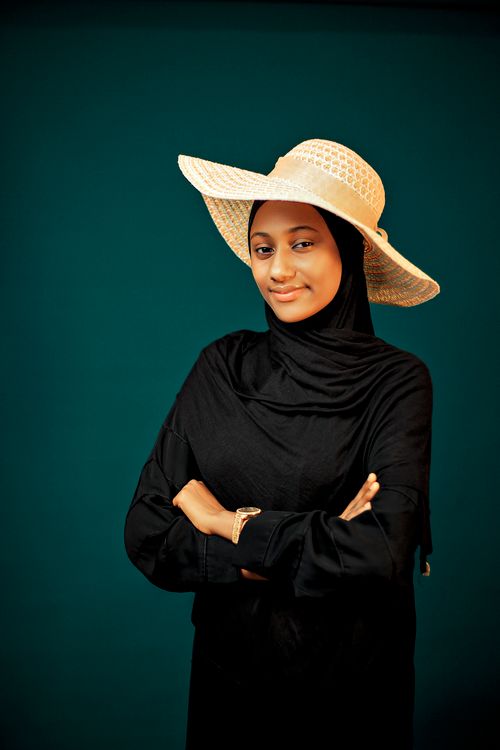 Free Portrait of a confident woman wearing a casual hat and a black hijab against a teal background. Stock Photo