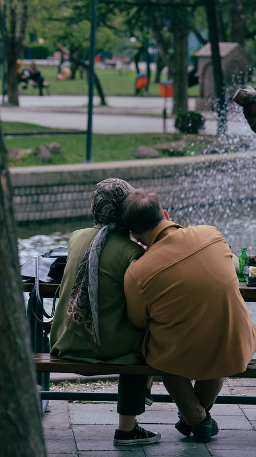 Free Older couple sitting by a fountain, sharing a peaceful moment in a city park. Stock Photo