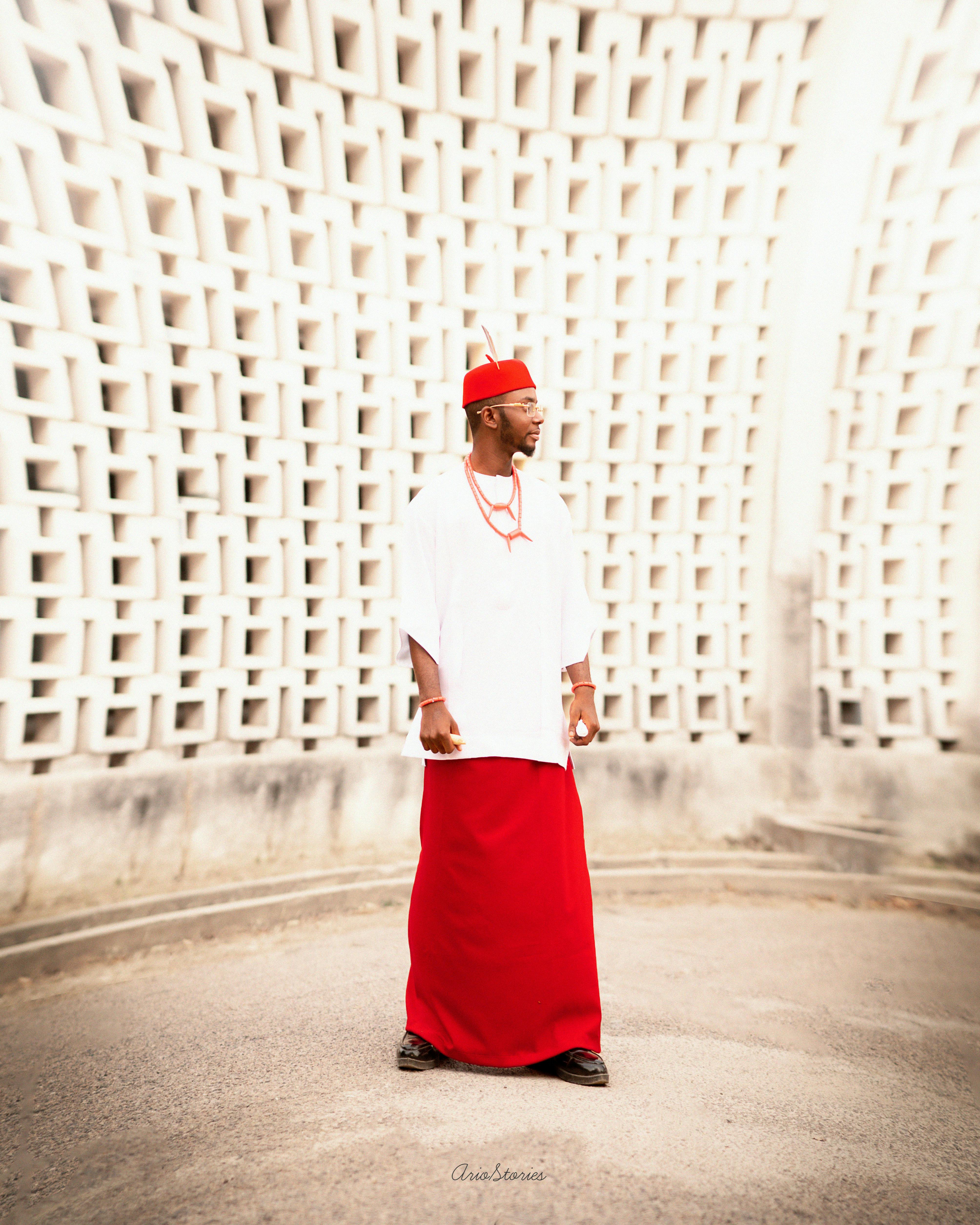 Man in traditional attire standing against a modern geometric wall outdoors.