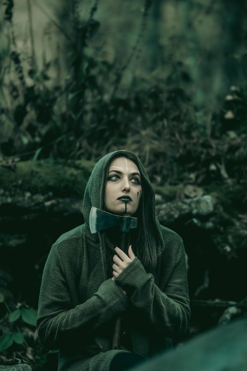 Free A hooded woman with face paint holds an axe in a dark, moody forest setting, embodying mystery. Stock Photo