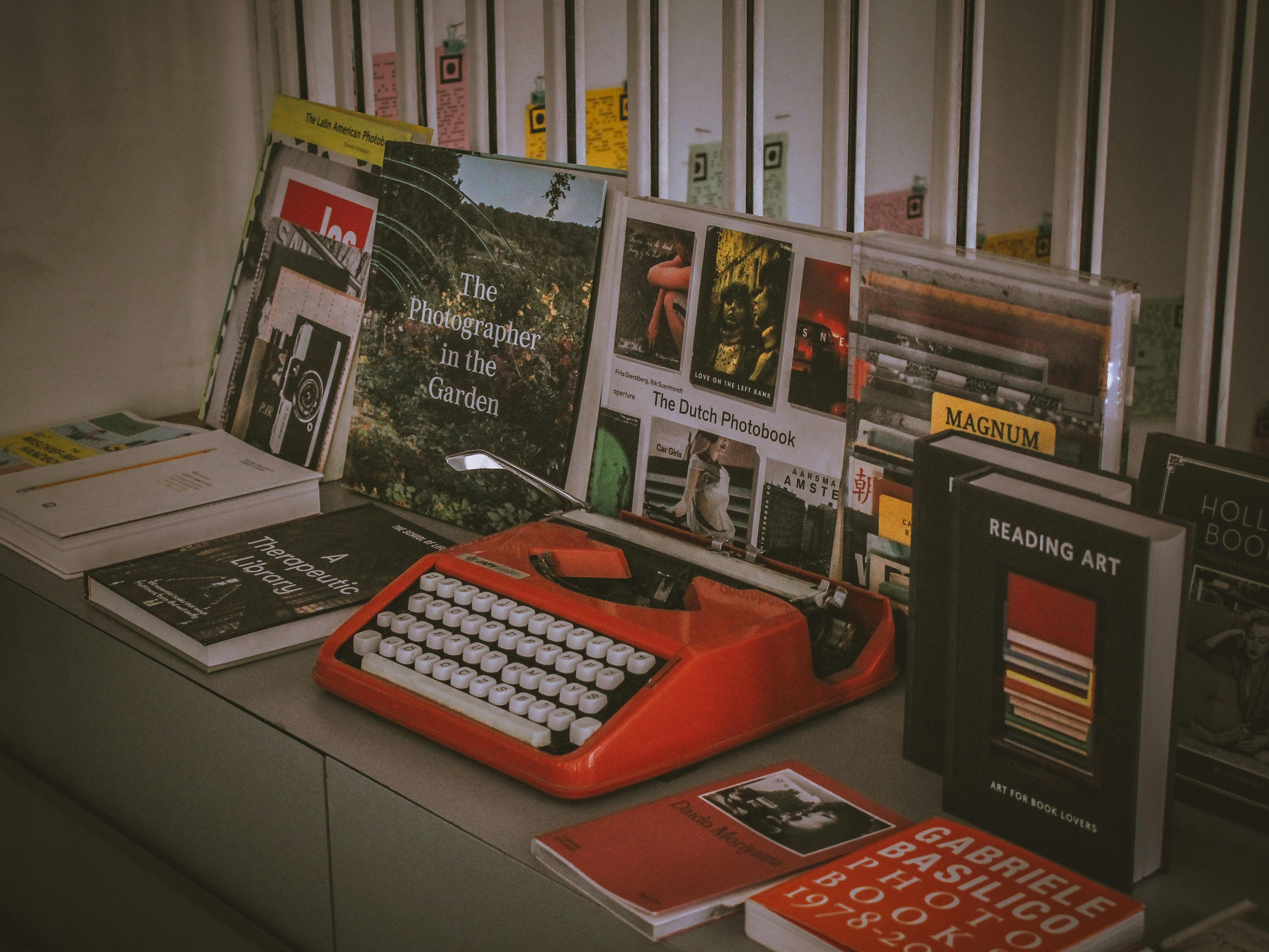 Kostenlos Eine leuchtend rote Vintage-Schreibmaschine, umgeben von einer Sammlung von Fotobüchern und Literatur in einem künstlerischen Arrangement. Stock-Foto