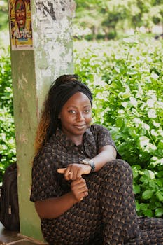 Young woman sitting by a pillar outdoors, surrounded by greenery and a festival poster.