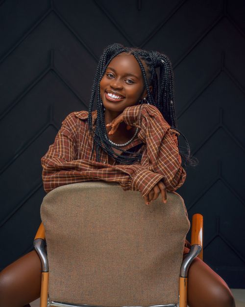 Free Portrait of a young woman with braided hair and a plaid shirt sitting happily on a chair indoors. Stock Photo