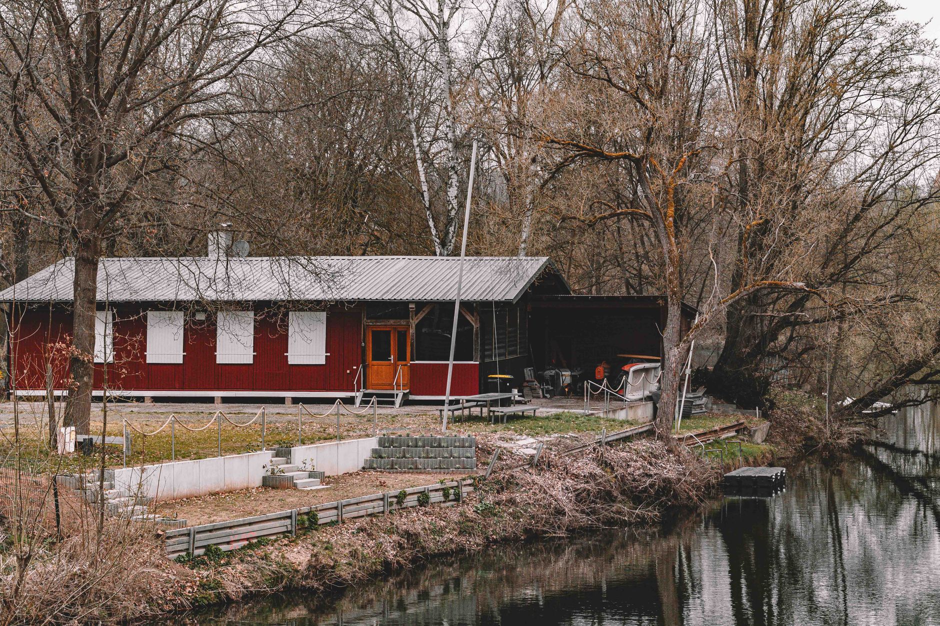 A serene riverside view of a rustic red cabin surrounded by bare trees in early spring, perfect for nature lovers.