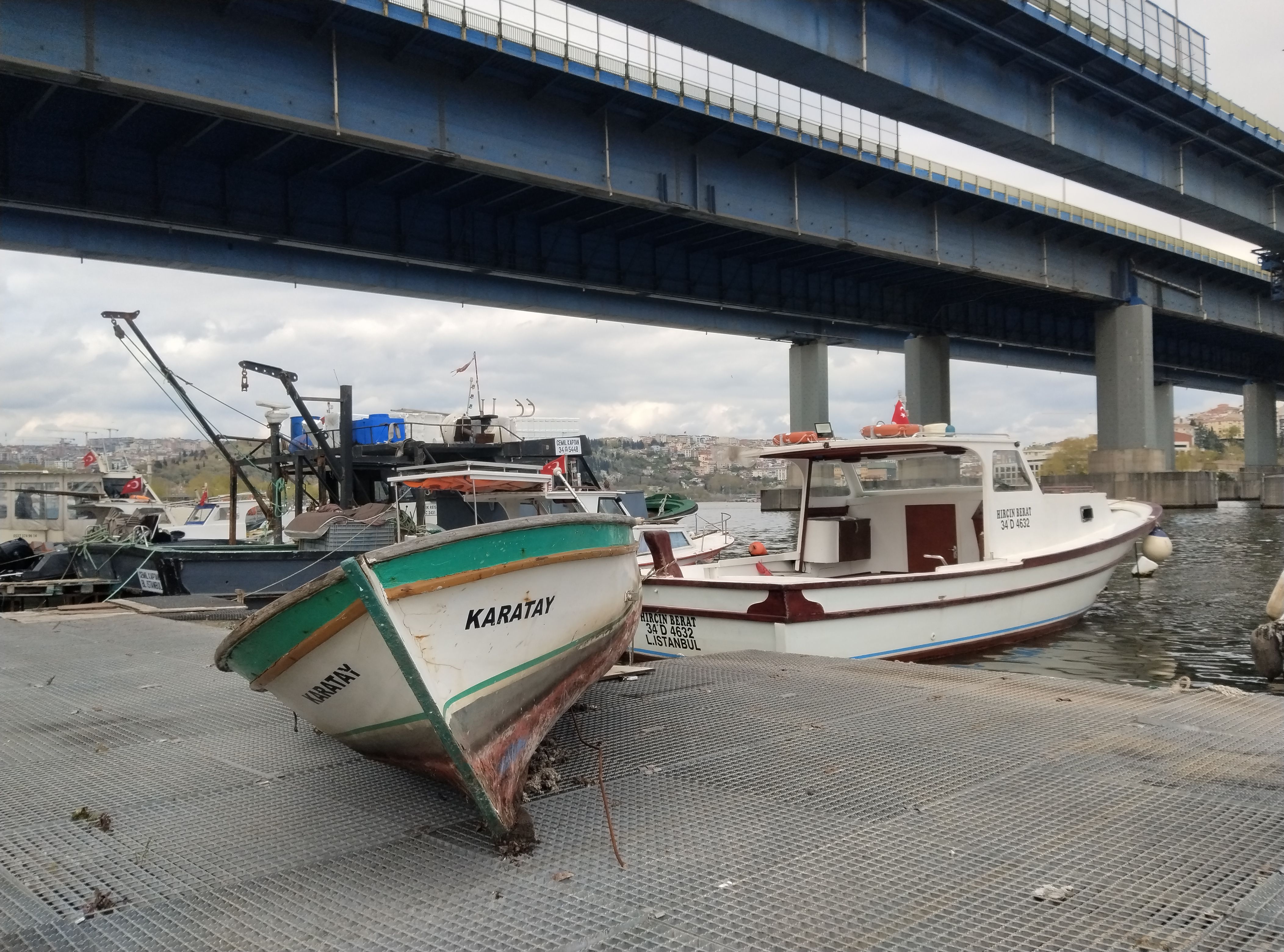 Two boats docked under a large bridge in Istanbul, Turkey, on a cloudy day.