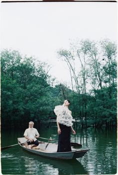 A stylish photoshoot featuring an elegant woman and an elderly man in a boat on a peaceful lake.