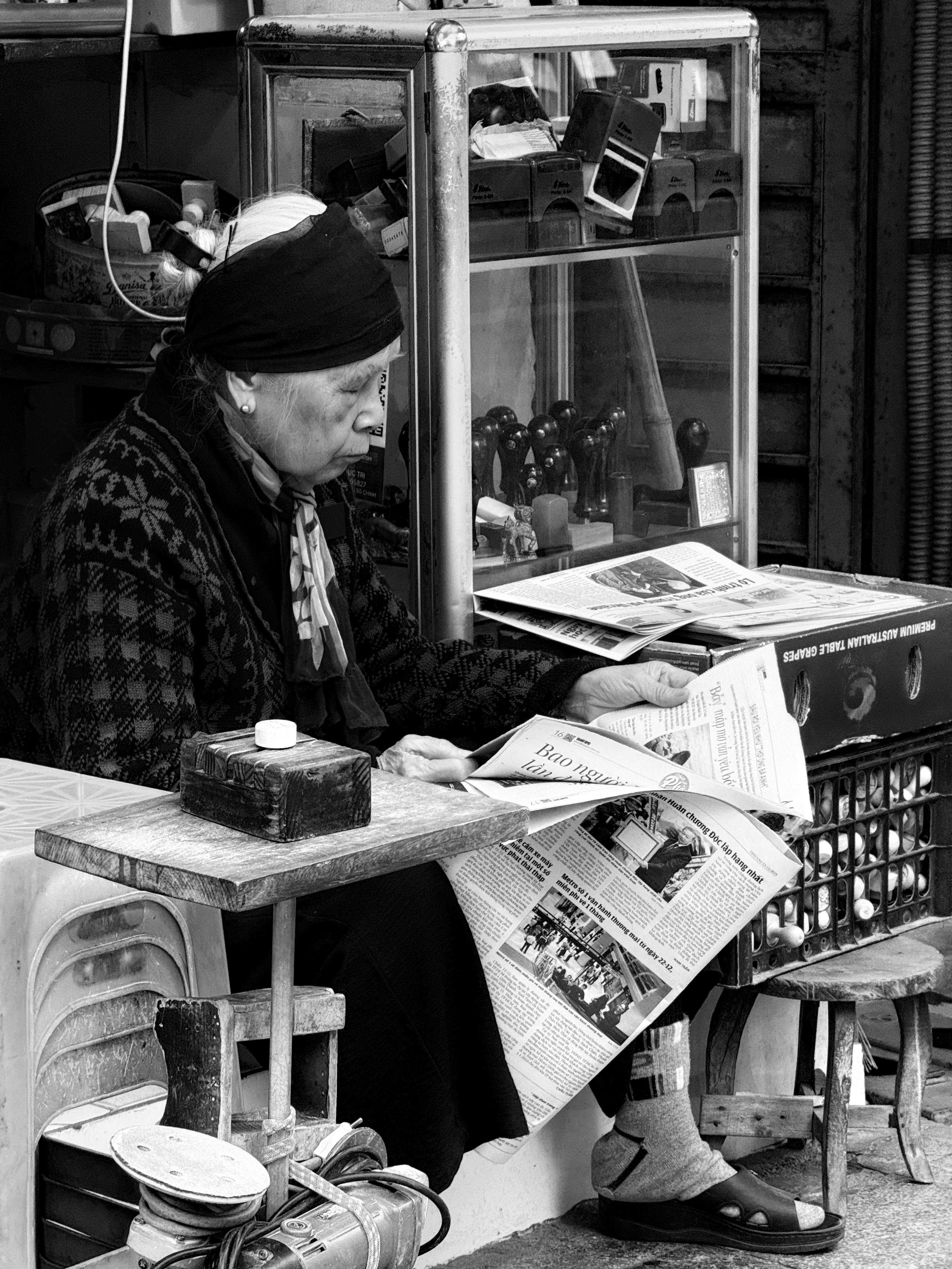 Senior woman sitting and reading a newspaper at an outdoor street market, showcasing a daily life scene.