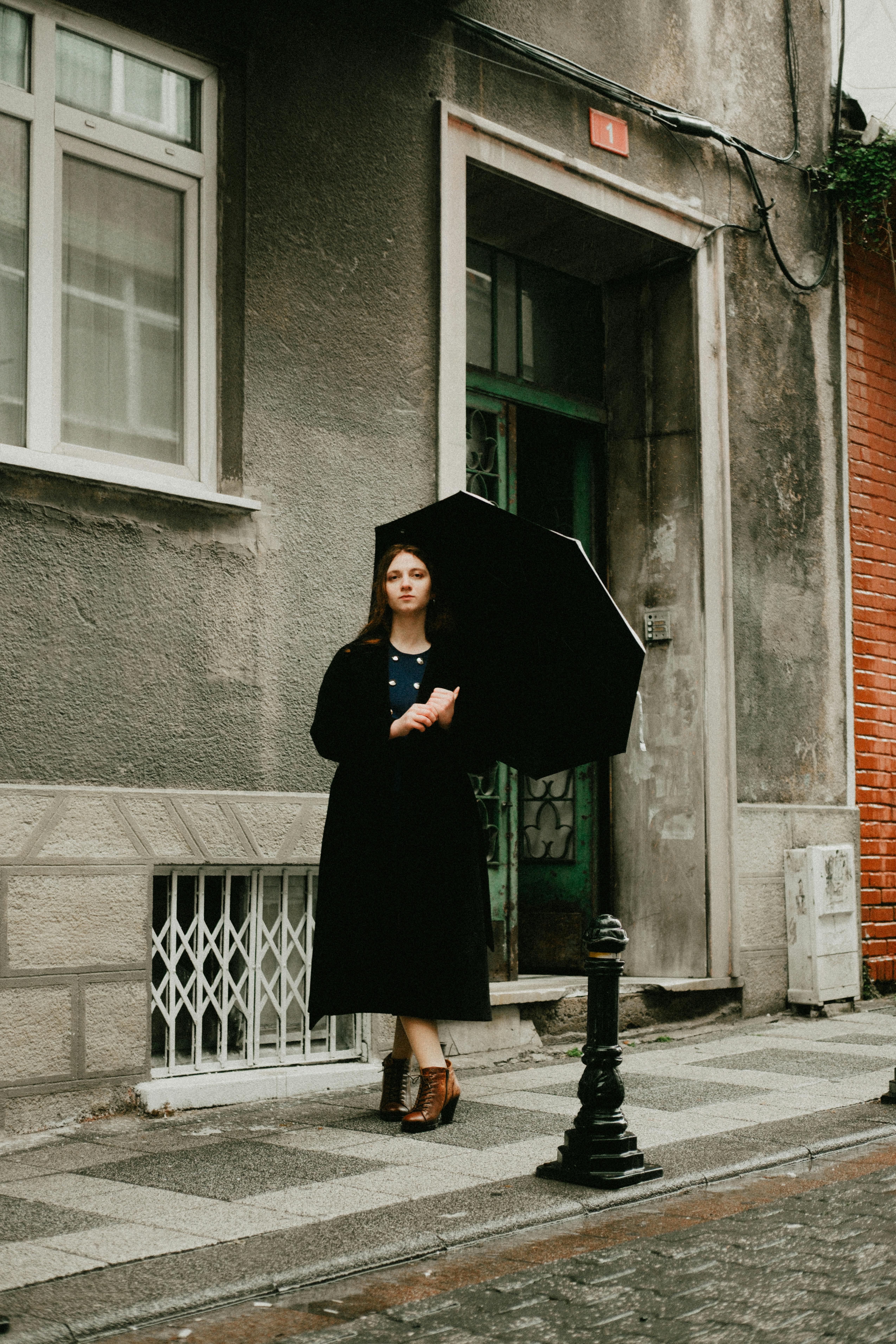 Woman in black coat with umbrella stands on a city street corner.