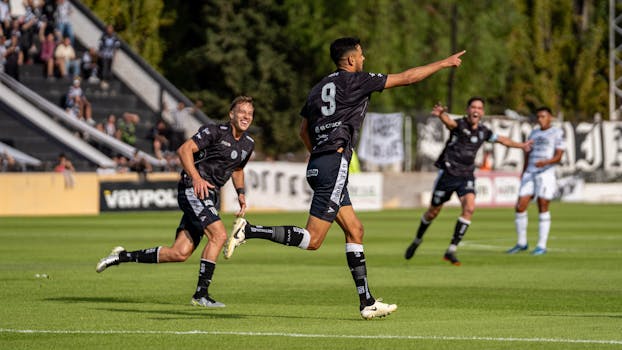 Football players in black jerseys celebrate a winning goal on a sunny outdoor field.