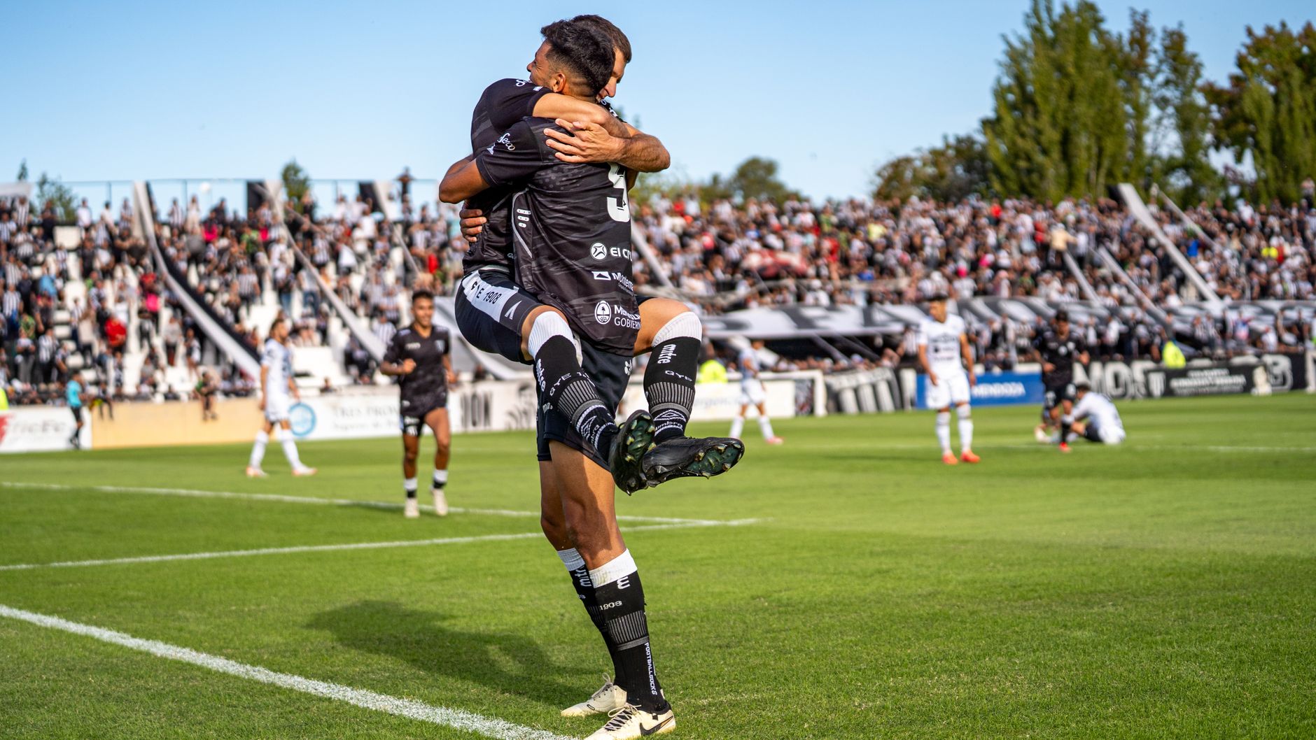 Two soccer players joyfully celebrate a goal during a match on a sunny day.