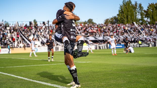 Two soccer players joyfully celebrate a goal during a match on a sunny day.