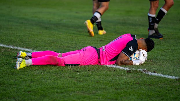 Soccer goalie in bright pink kit lying on grass field during a match.