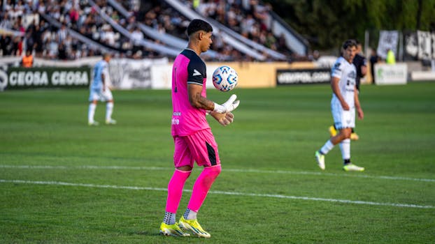 A soccer goalkeeper in pink uniform catches a ball during a match on a grassy field.