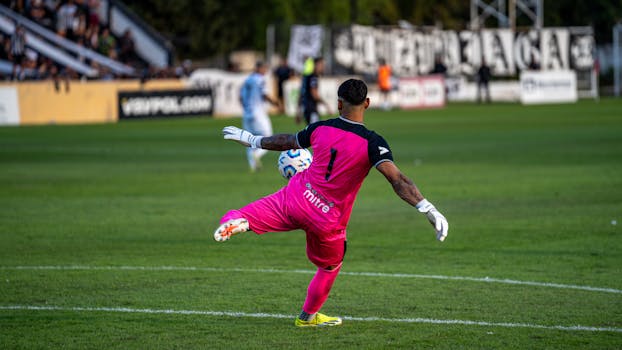 A goalkeeper in bright pink kit kicking a soccer ball during a match.