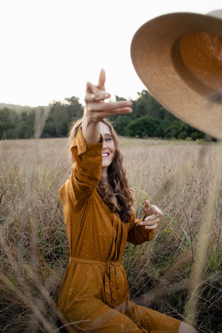 A woman in a yellow dress reaches for a straw hat in a sunlit meadow.