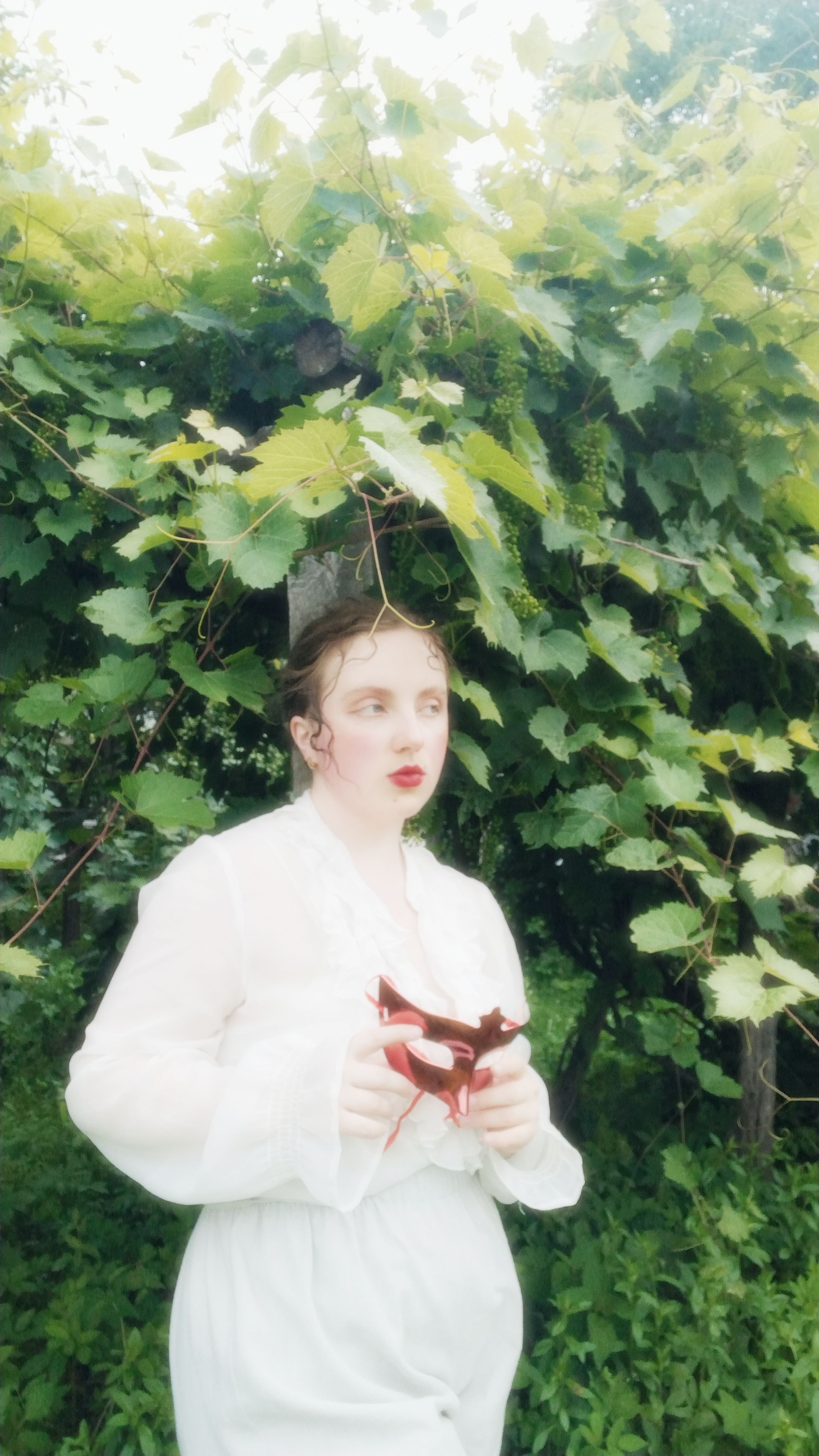 Artistic portrait of a woman in white holding a red object under lush green foliage.