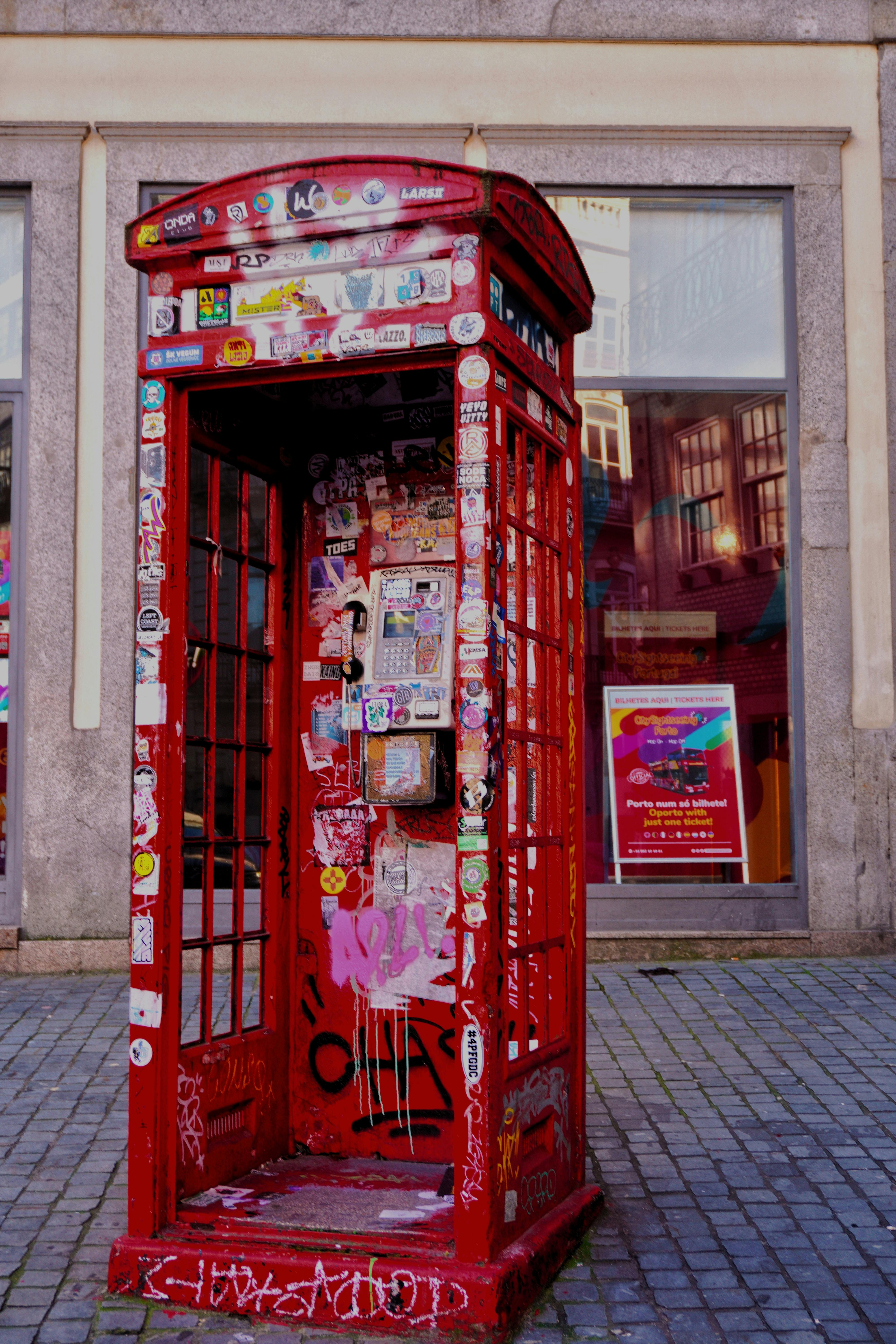 A red phone booth covered in graffiti and stickers in an urban setting.