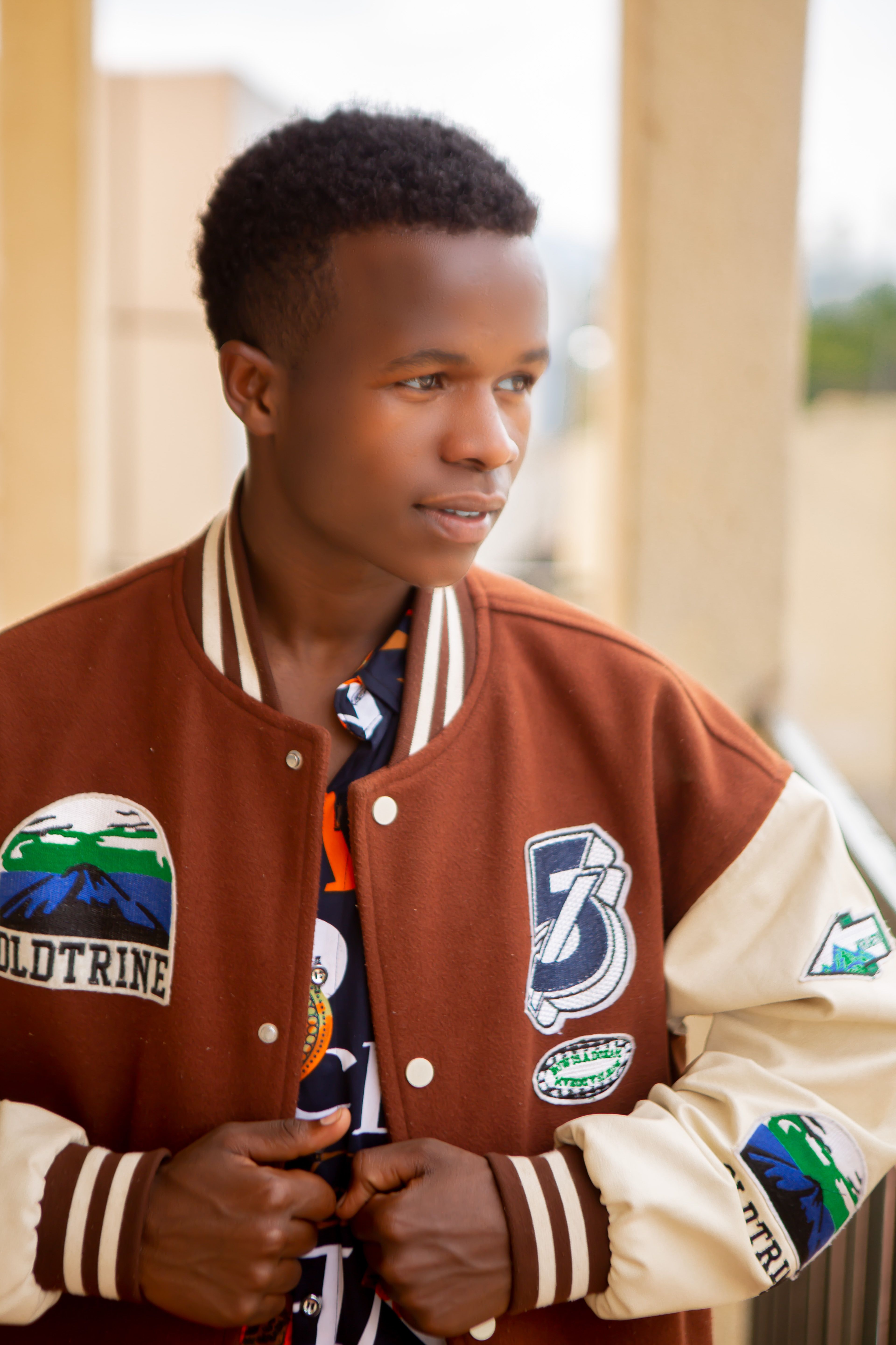 Young man wearing a brown varsity jacket stands outdoors with casual and thoughtful demeanor.