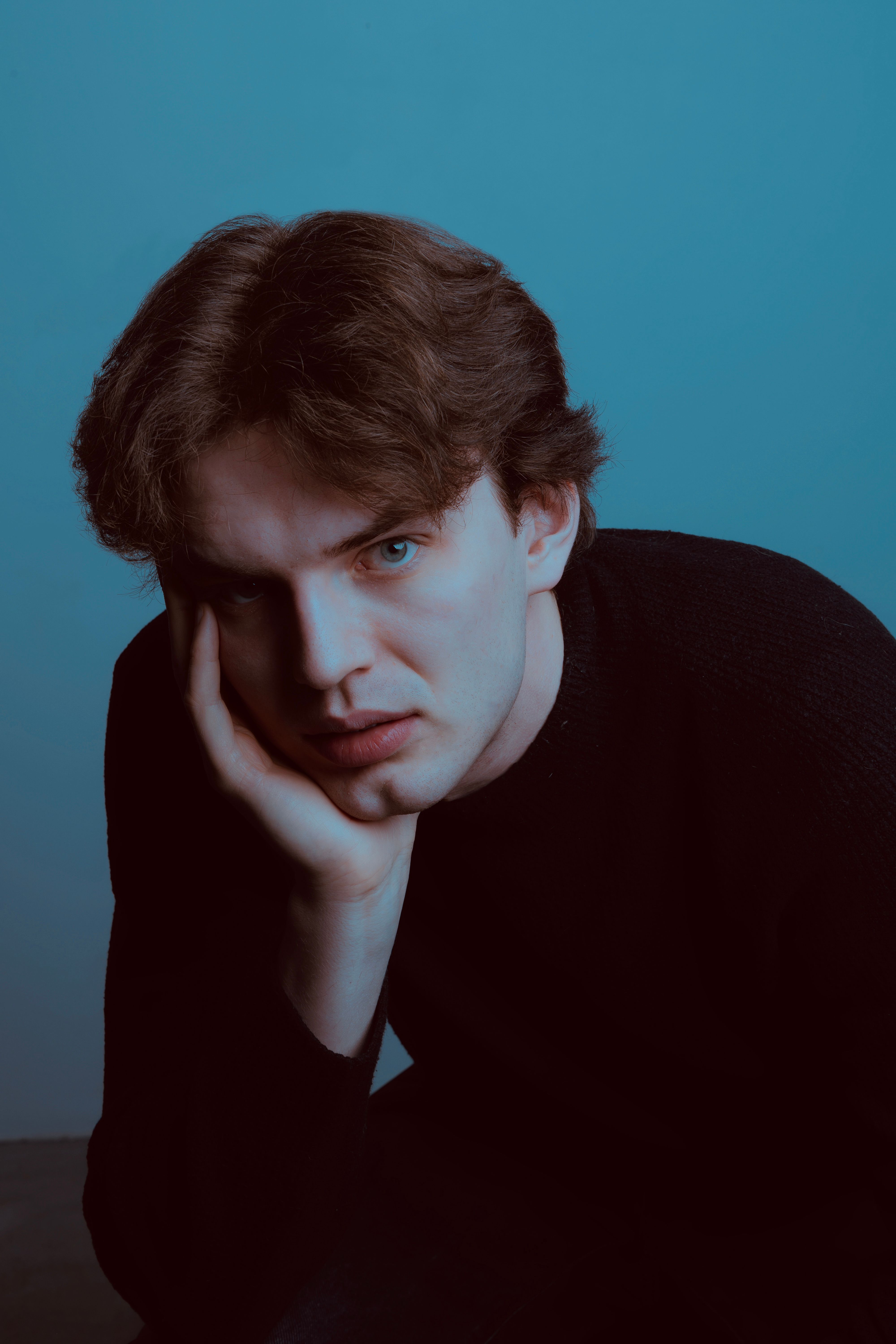 A thoughtful young man in a dark sweater against a blue backdrop, conveying introspection.