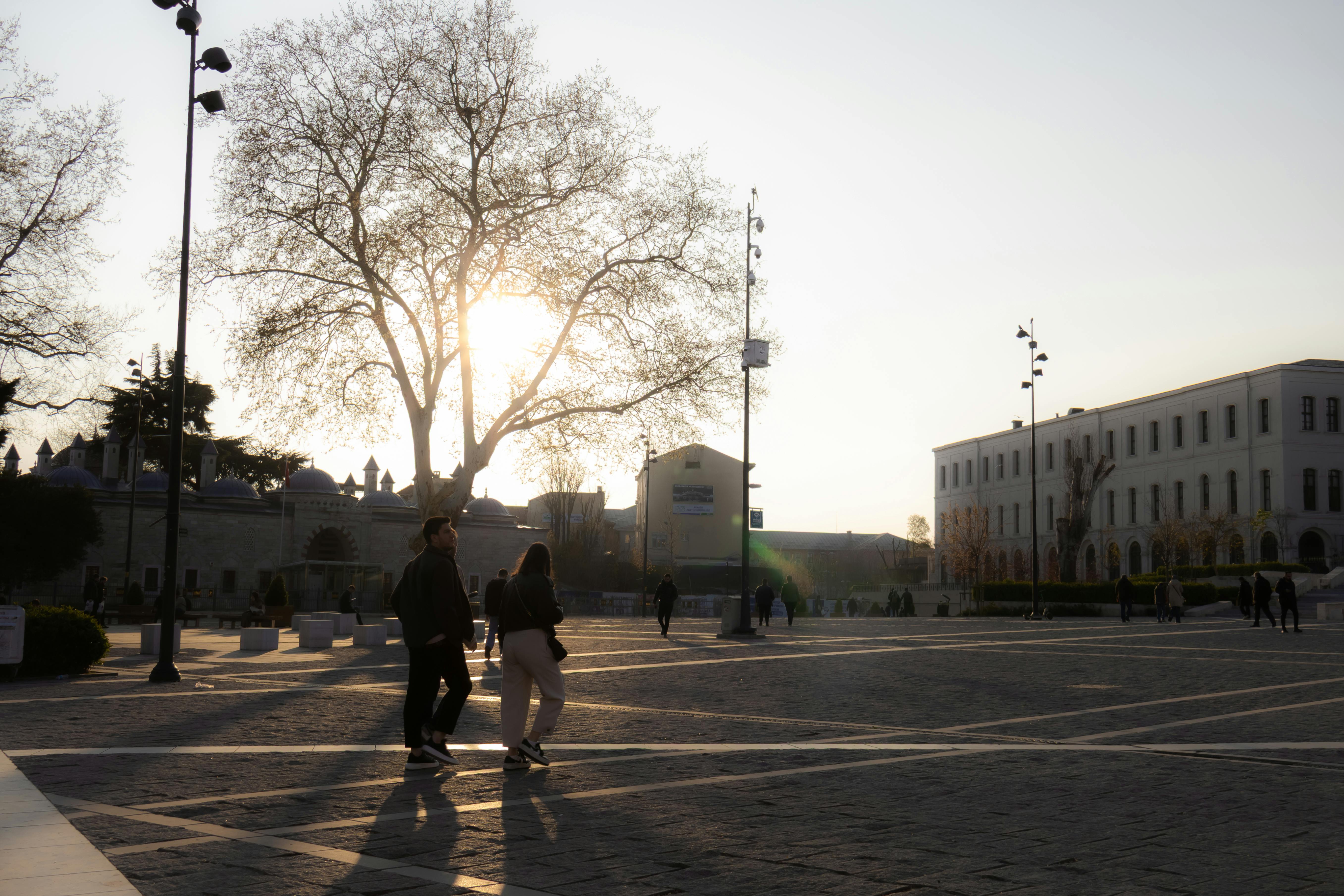 People walking at sunset in a historic square in Istanbul, Türkiye, casting long shadows.