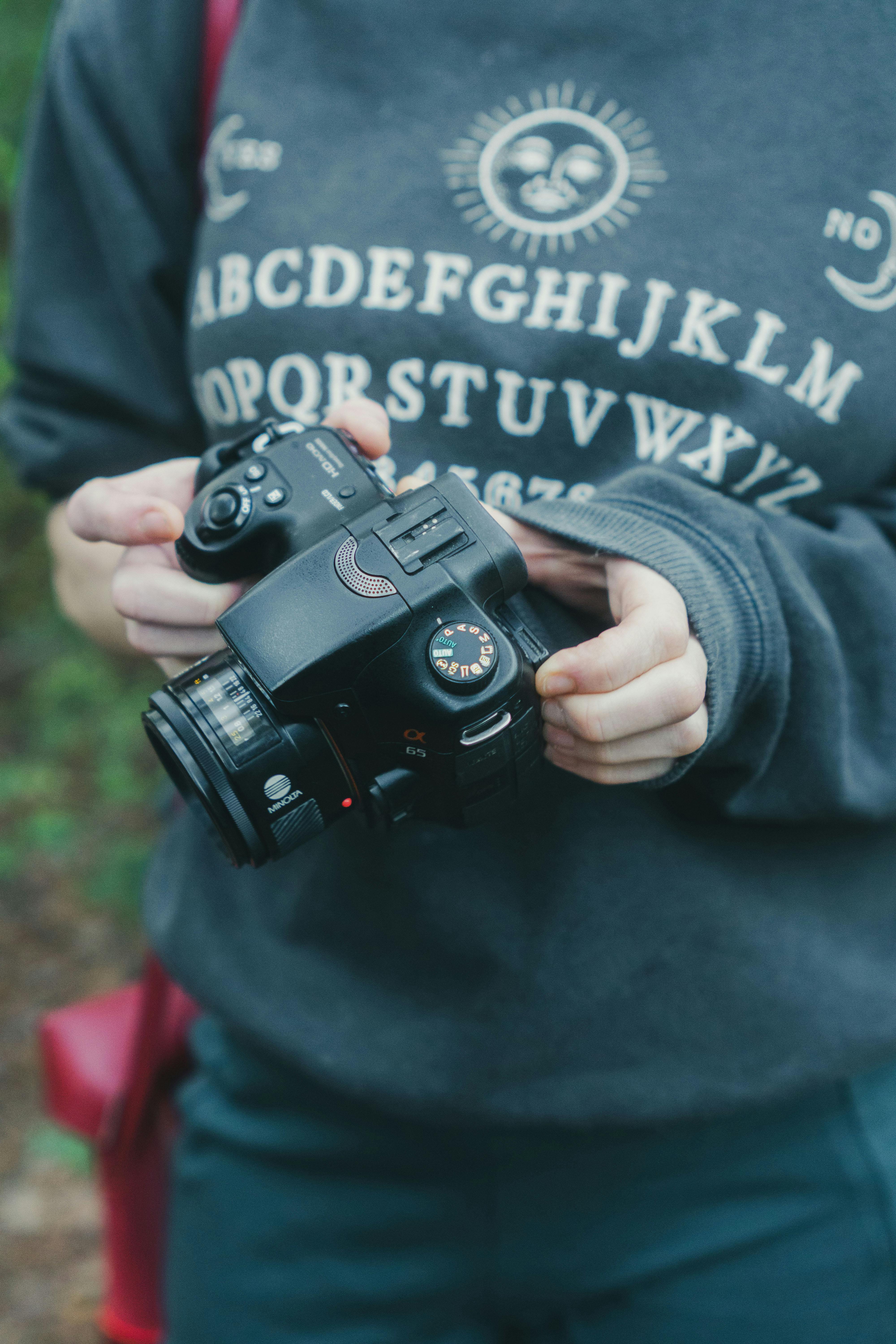 Free A young adult wearing a graphic sweater holds a DSLR camera outdoors in Houston, Texas. Stock Photo