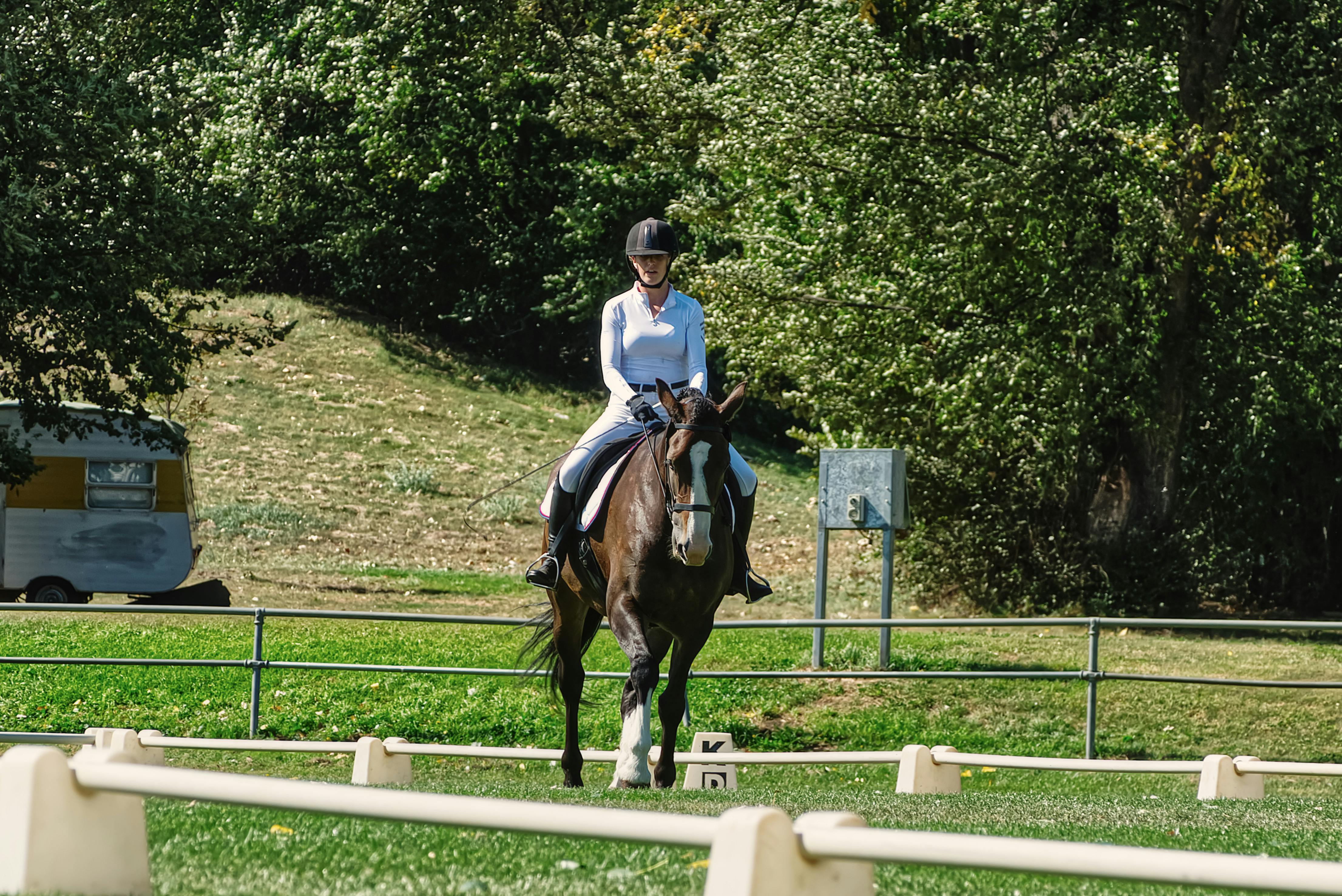 A woman rides a horse in an outdoor dressage arena surrounded by greenery.