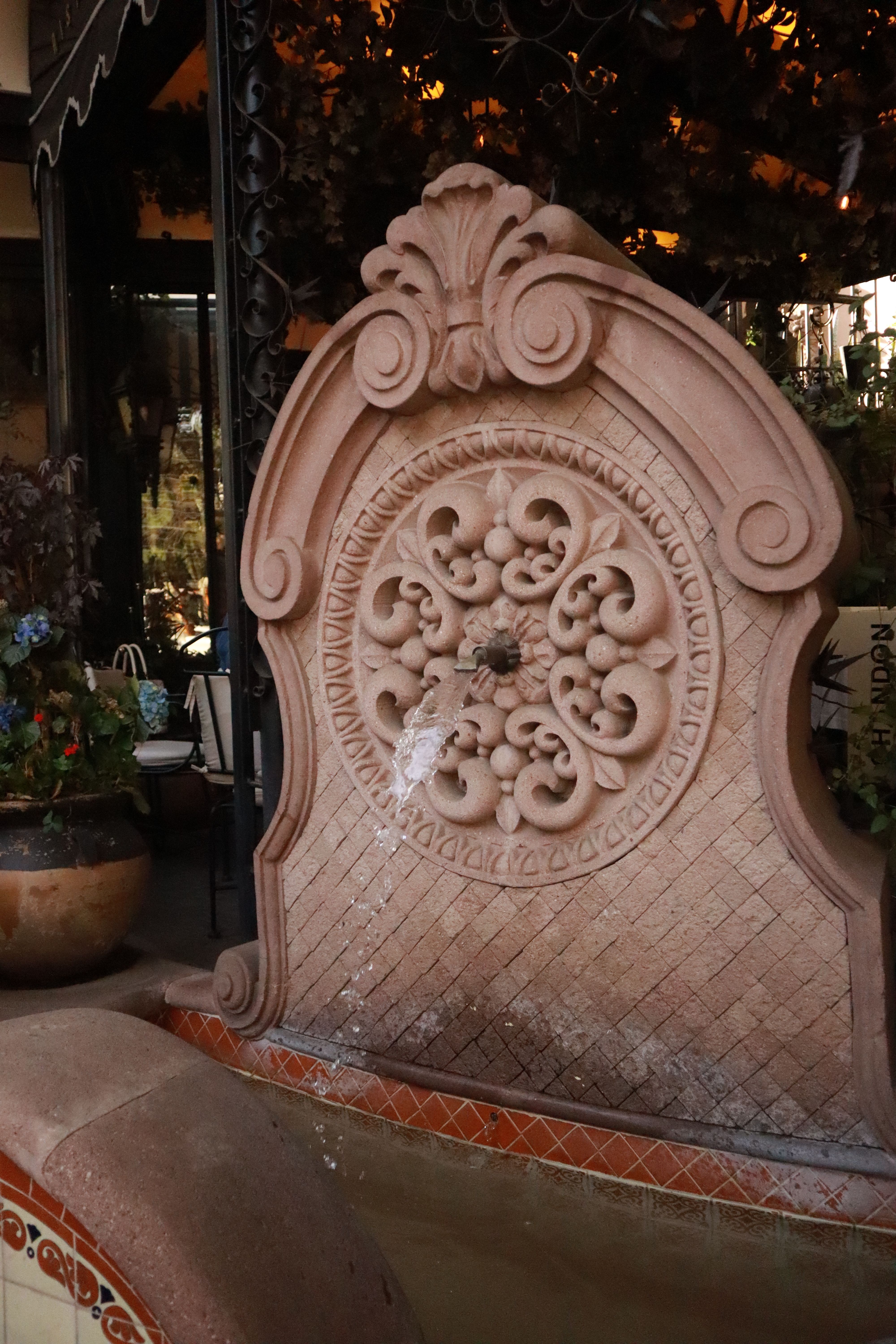 Side patio design with a small wall-mounted stone water fountain attached to the side fence, with potted ferns around the base