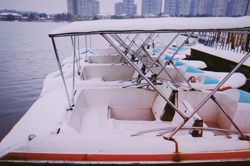 Free A serene winter scene of pedal boats covered in snow by a lakeside with city skyline in the background. Stock Photo