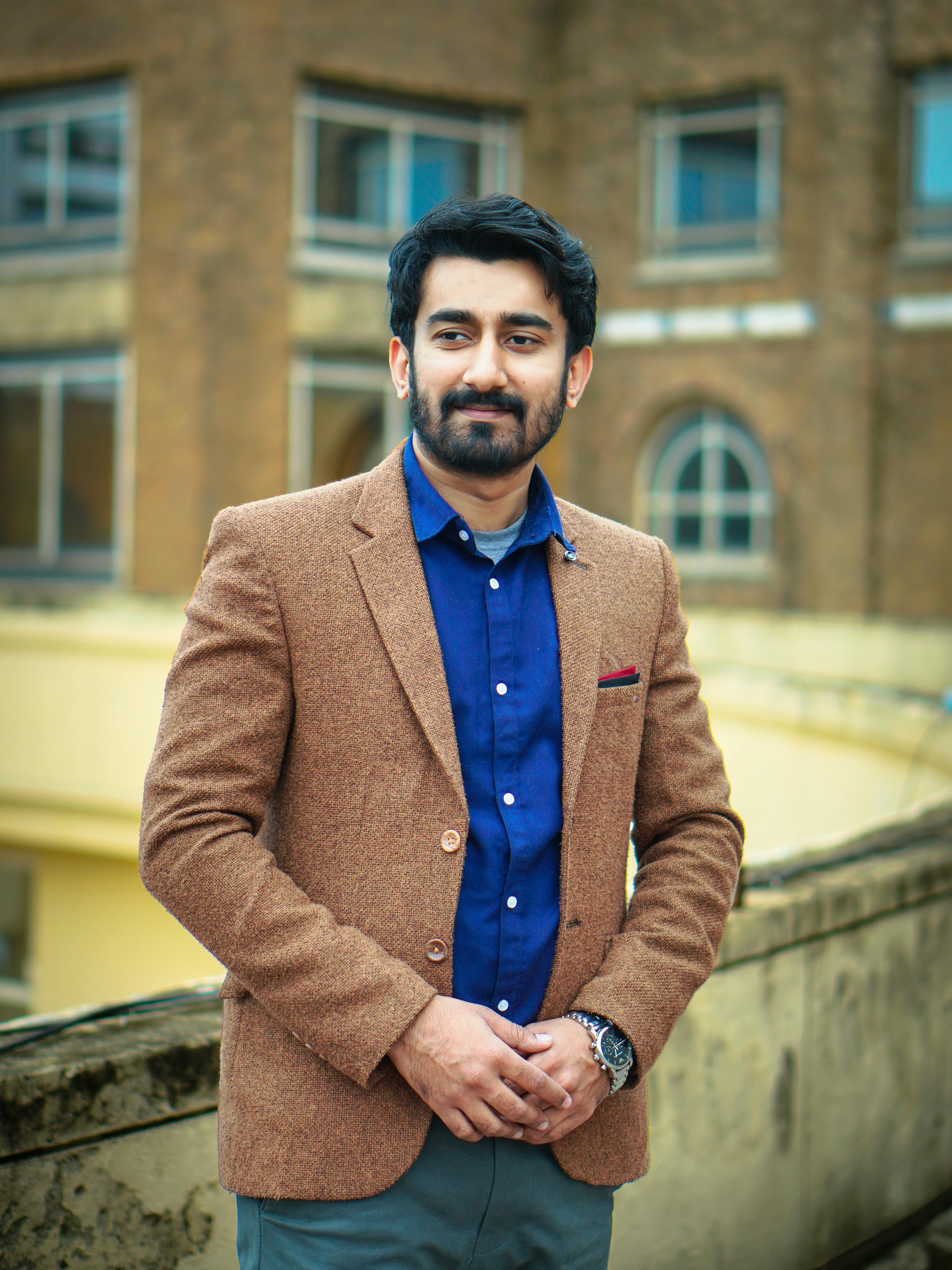 Confident man in brown blazer posing on a rooftop with a historical building in the background.