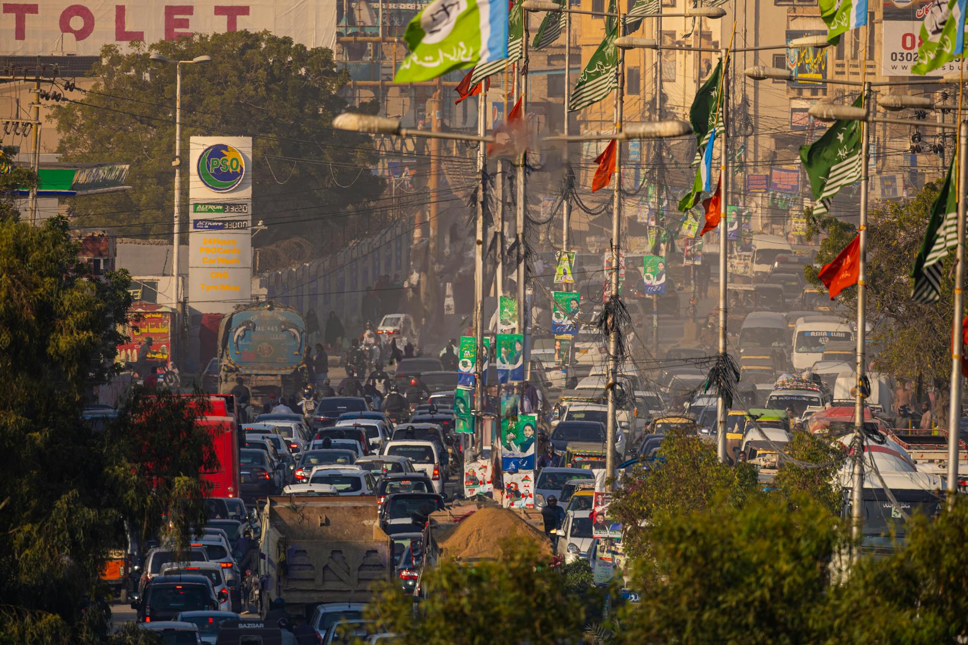 A busy Karachi street filled with vehicles, colorful flags, and political posters during rush hour traffic.