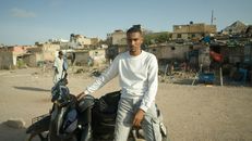 A young man sits on a motorcycle against a rustic urban backdrop in Morocco.