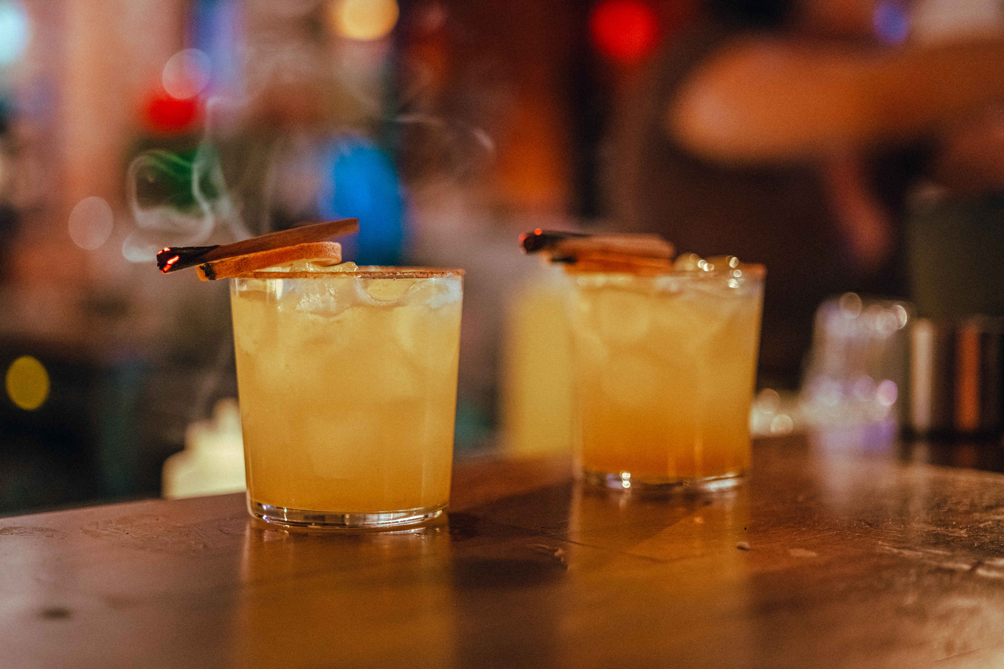 Two orange cocktails with cinnamon sticks on a wooden bar in a cozy, dim pub.