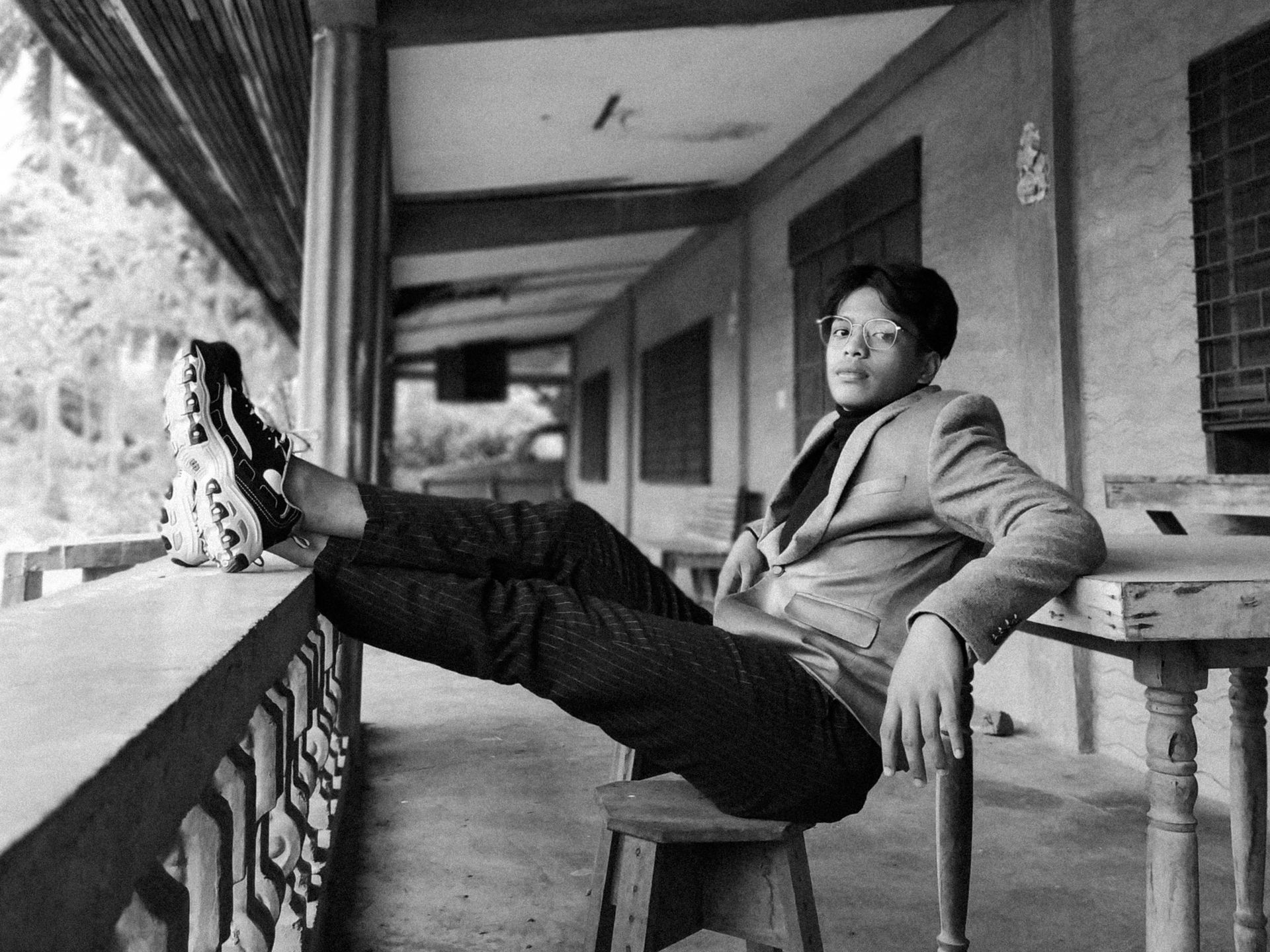 Black and white portrait of a young man reclining on a chair outdoors in a stylish outfit.