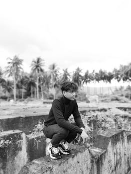 A young Asian man sitting outdoors with blurred palm trees in the background.