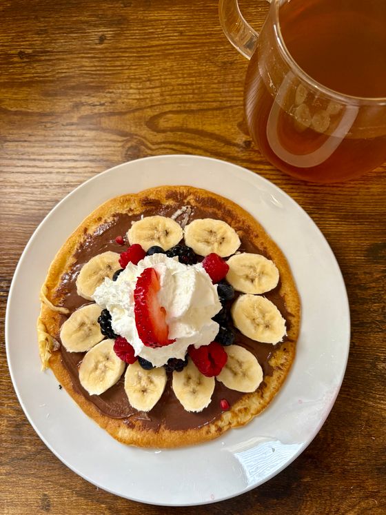 Large pancake spread with chocolate hazelnut, ringed with banana slices, berries and whipped cream