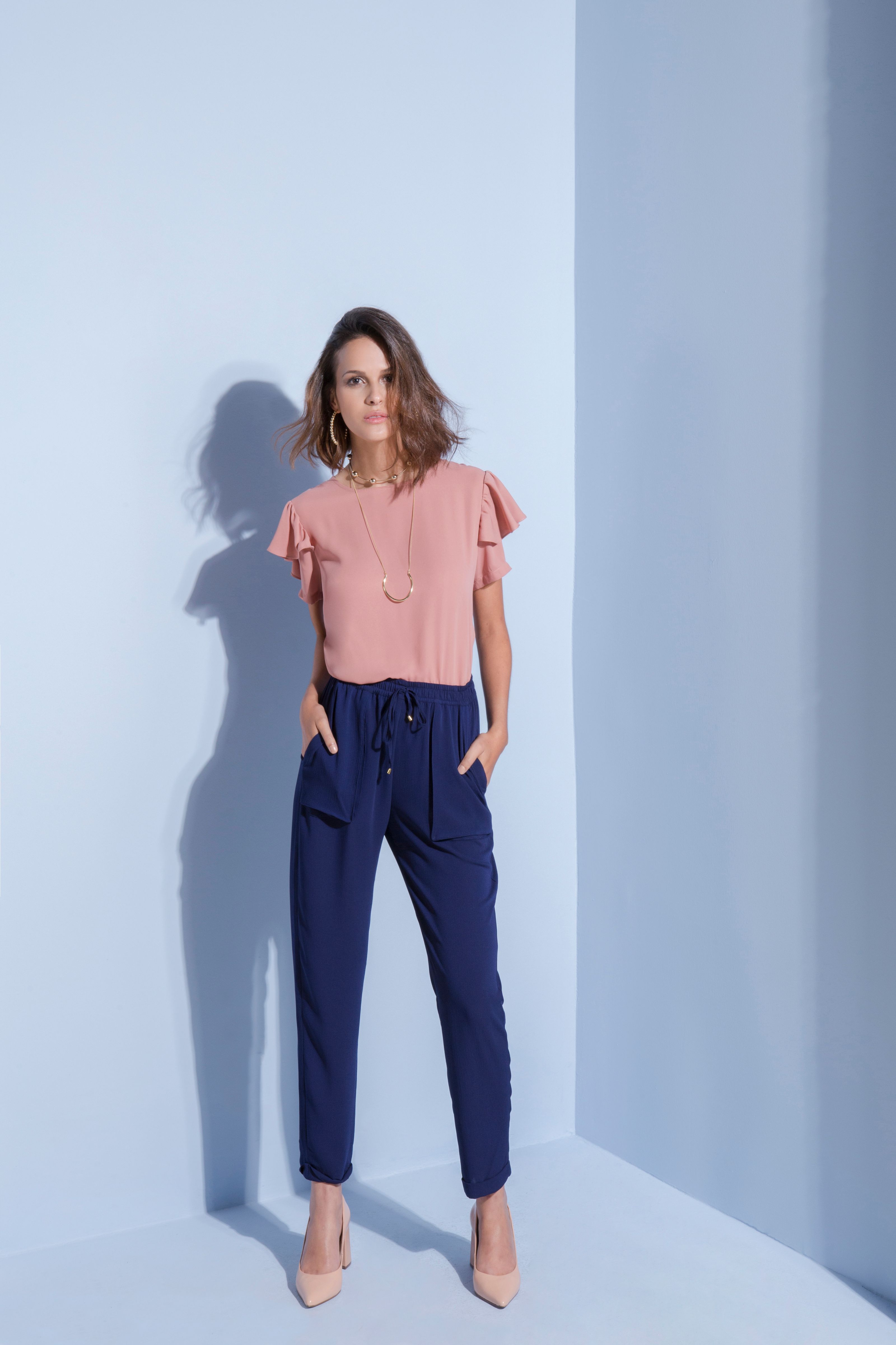 Mulher elegante posando com blusa rosa e calça azul-marinho sobre um fundo claro, personificando a moda contemporânea.