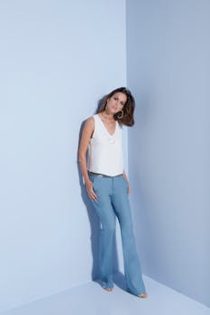 Stylish woman in fashionable blue pants and white top posing elegantly indoors on a light blue background.
