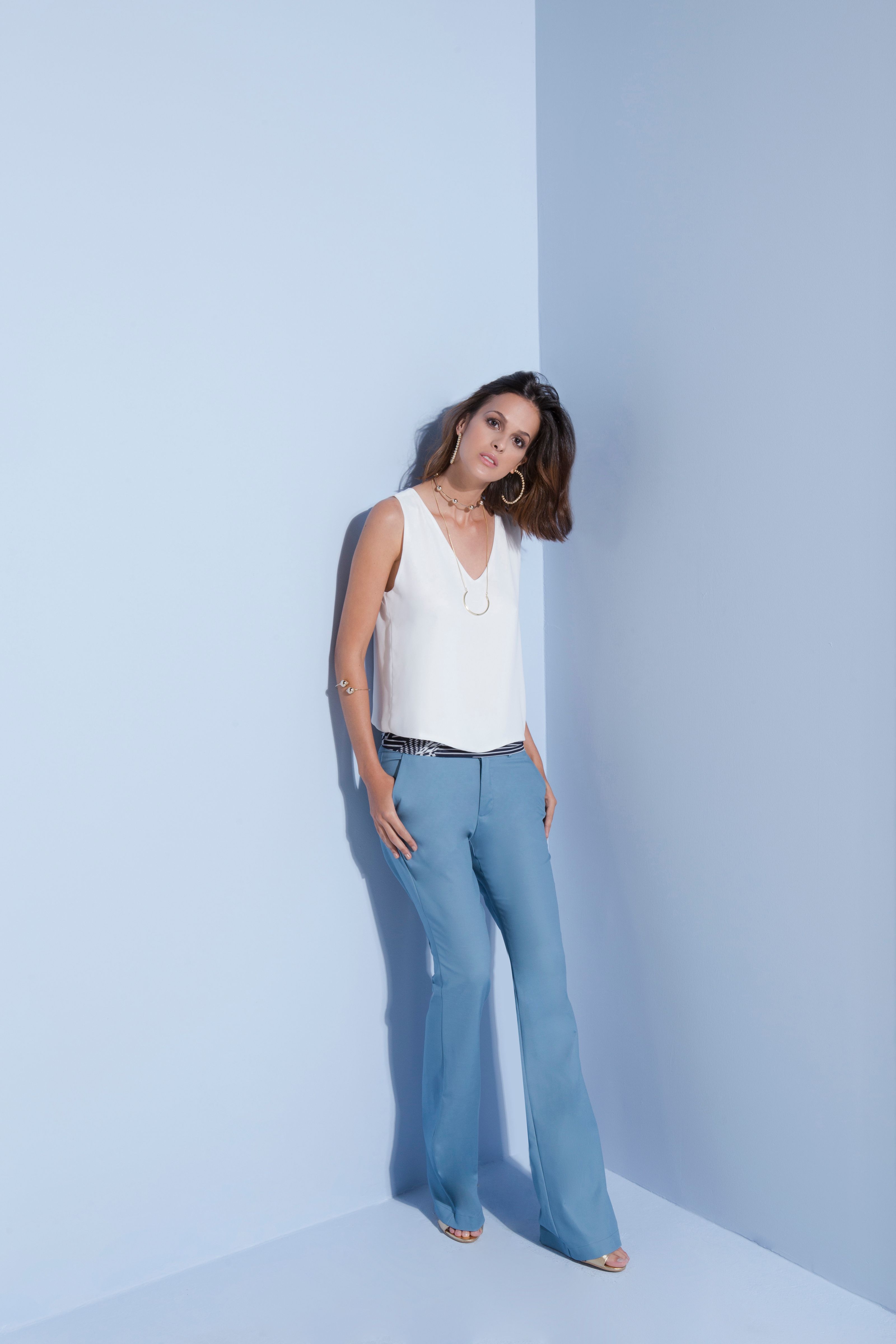 Mulher estilosa com calça azul e blusa branca posando elegantemente em um ambiente interno sobre um fundo azul claro.