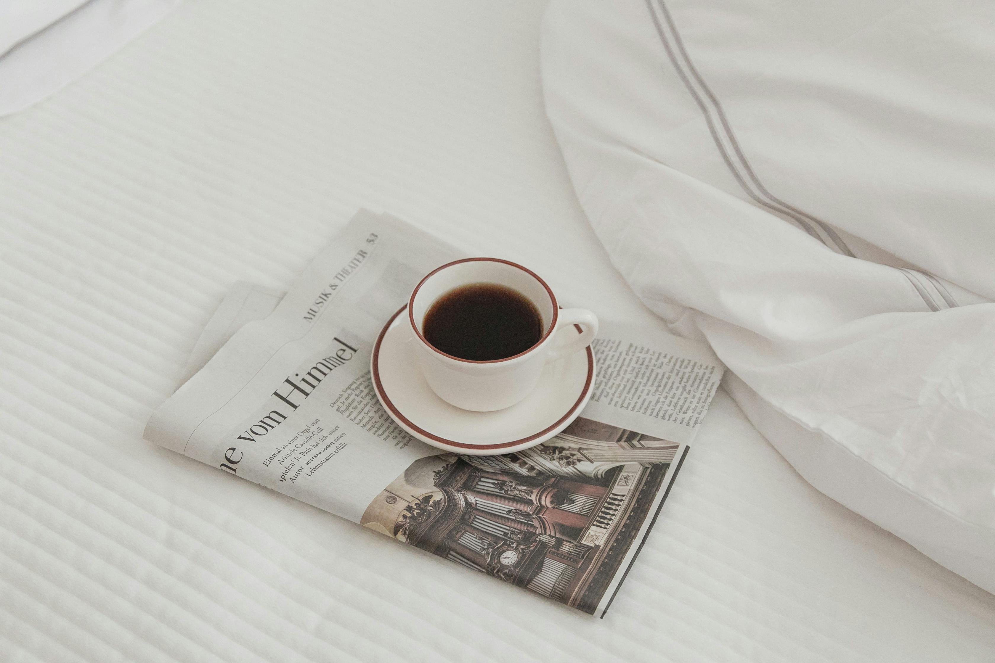Minimalist morning scene with a cup of coffee and newspaper on a neatly made bed.