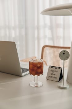 Cozy workspace featuring a laptop, iced coffee, and motivational note in soft lighting.