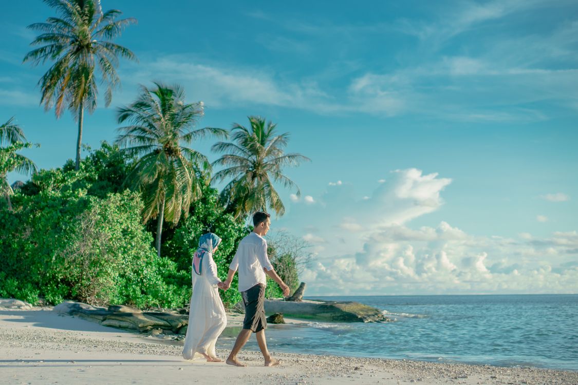 A couple holding hands while walking along a tropical beach lined with palm trees