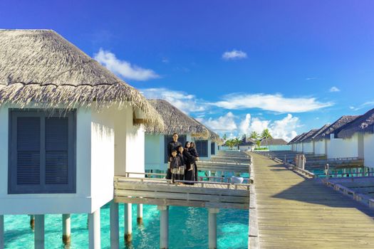 Family enjoys a sunny day on a resort's boardwalk with overwater bungalows and clear blue sky.